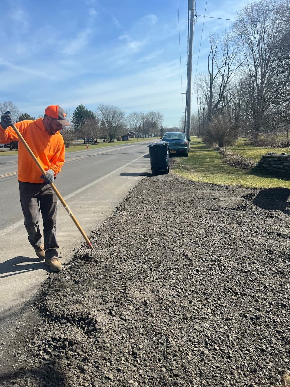 Worker in orange safety shirt raking gravel on roadside during excavation work
