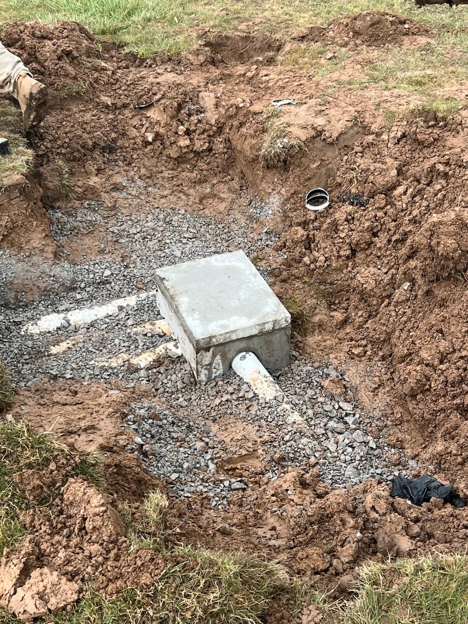 Concrete junction box with white pipe on gravel base in excavated area