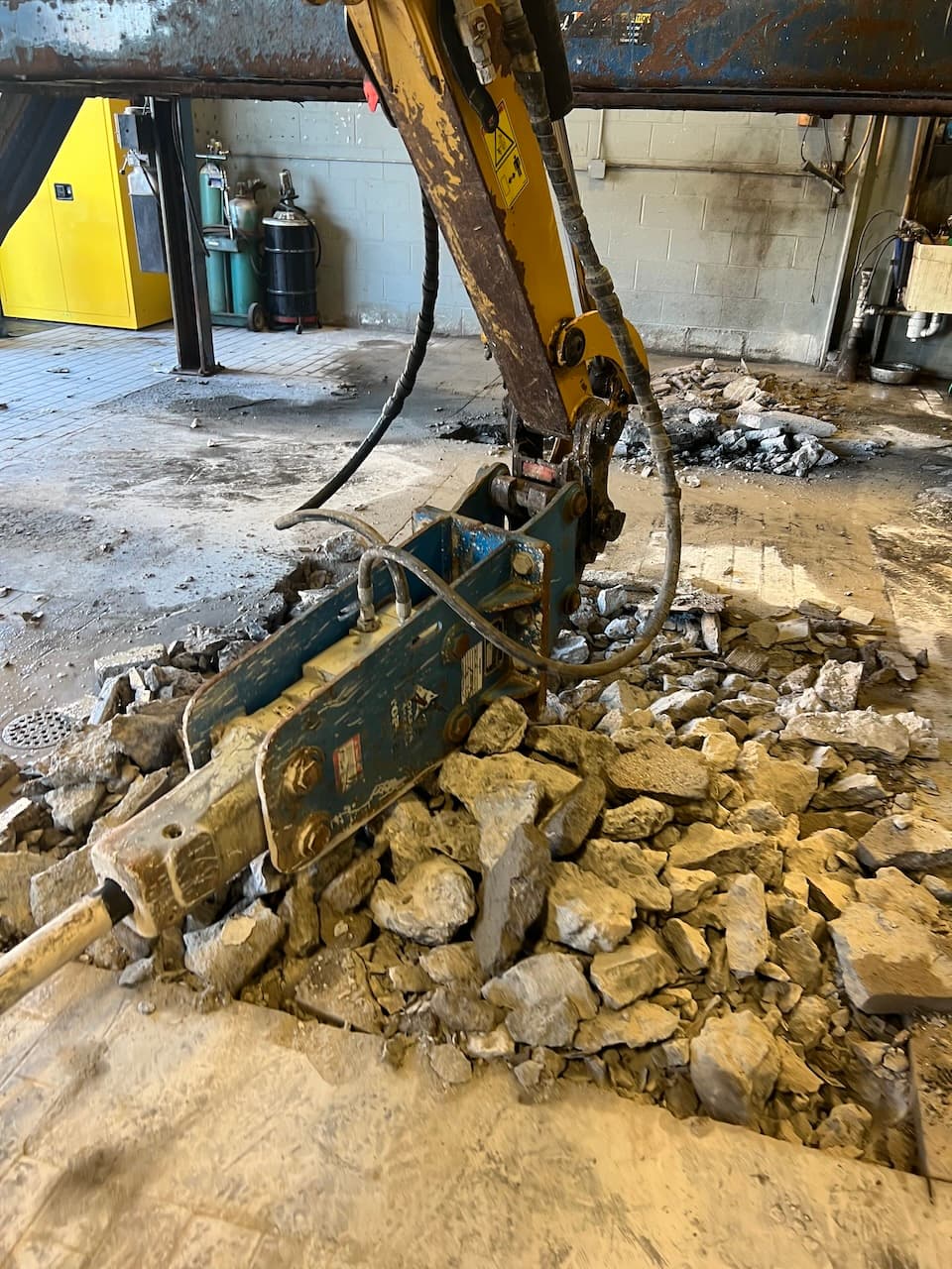 Yellow excavator breaking up concrete floor inside building during demolition