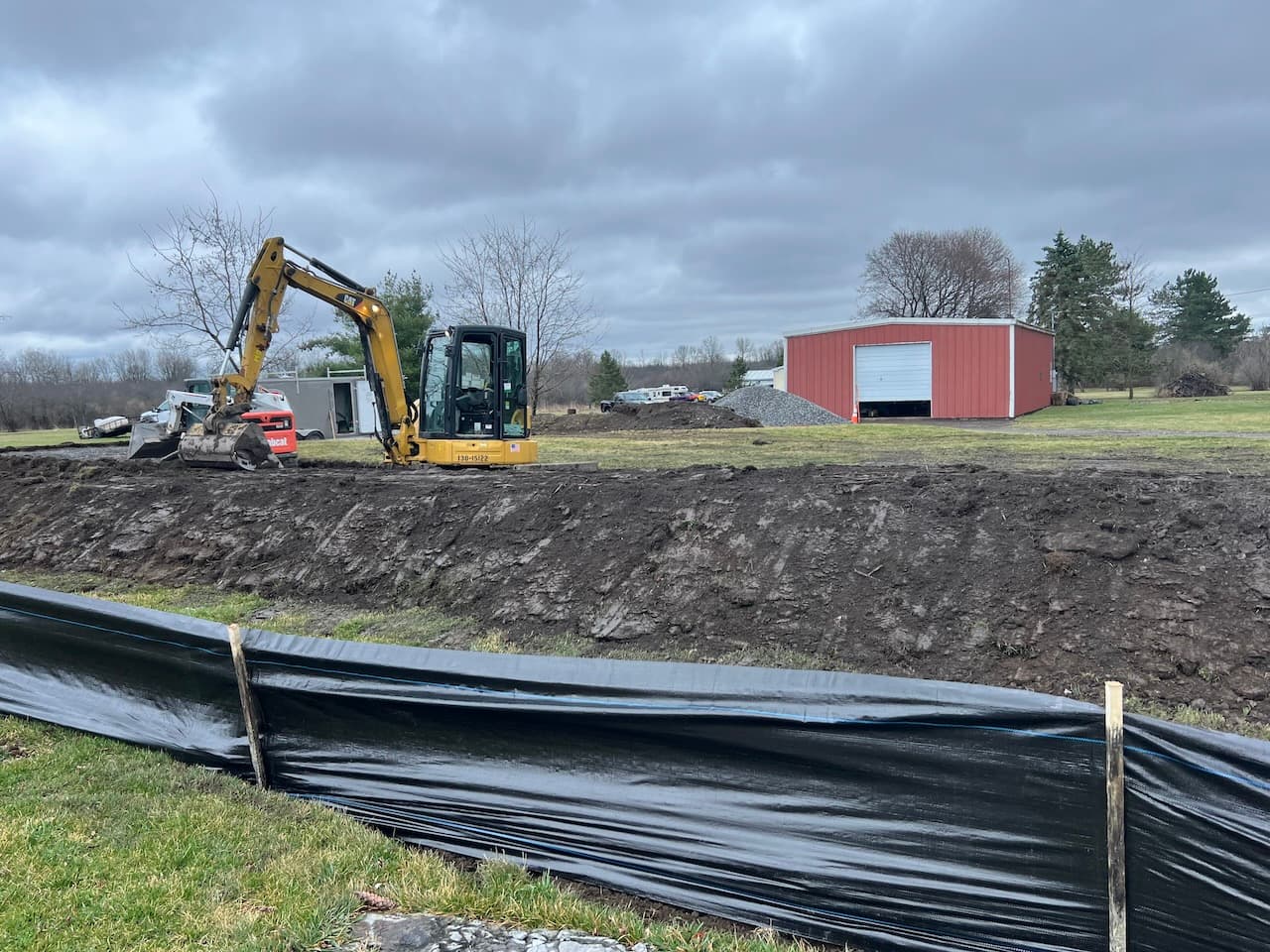 Yellow excavator preparing site for septic system installation with gravel pile and red building in background