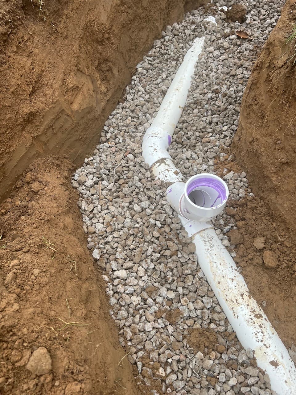 White PVC pipe with purple coupling laid in gravel bed trench