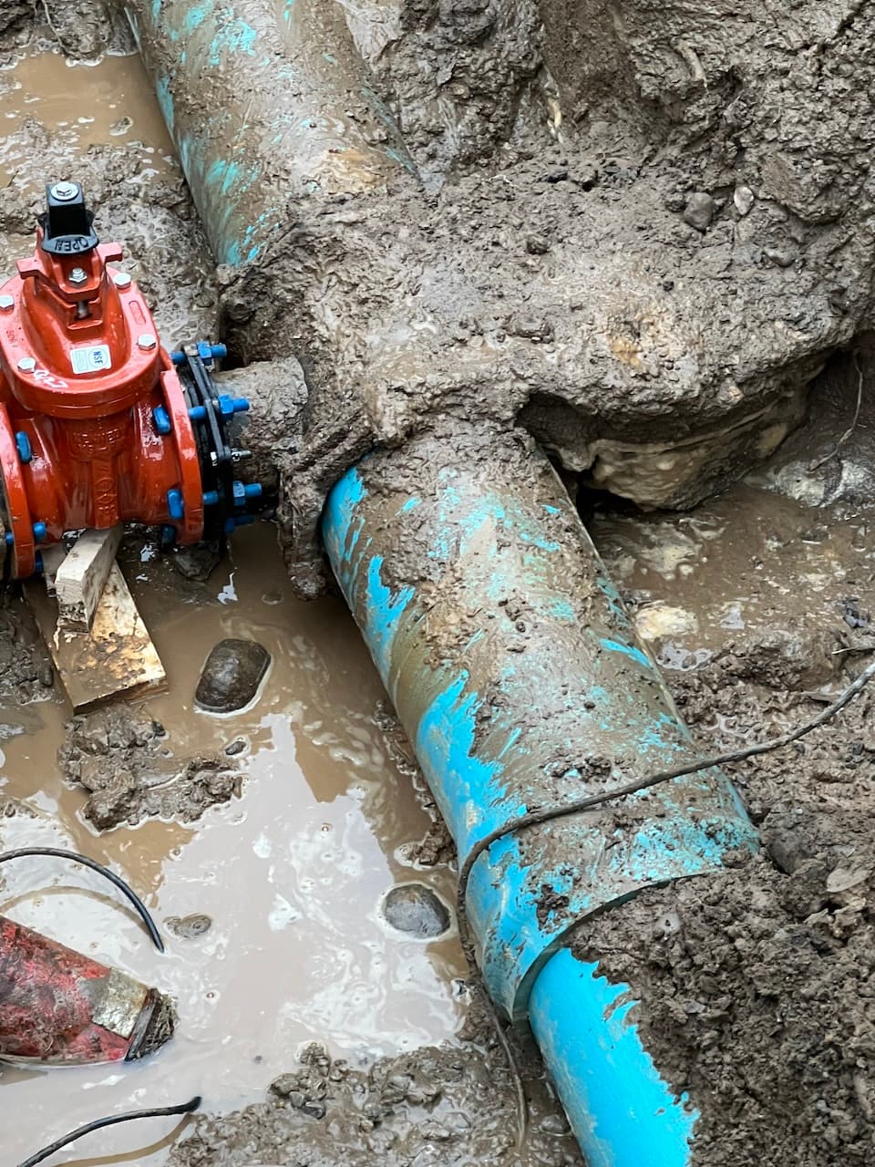Red gate valve connected to blue water main pipe in muddy trench